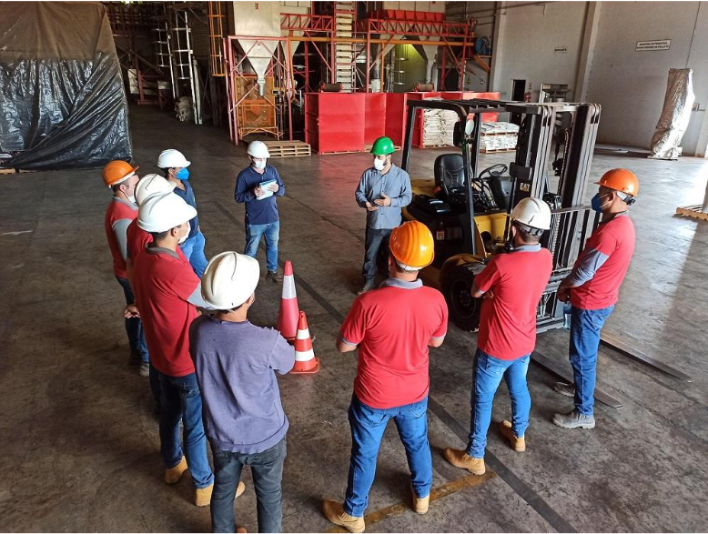 Warehouse safety briefing with a group of workers wearing hard hats and high-visibility gear standing around a forklift. The instructor, in a green hard hat, appears to be explaining safety procedures, with orange traffic cones marking the gathering area in the industrial setting.